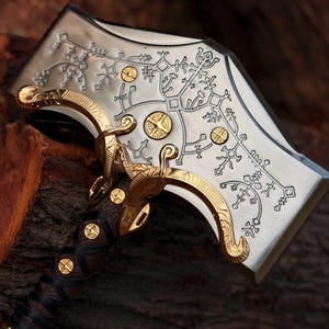 Pu&ograve; includere: Una testa d'ascia in metallo color argento con intricati motivi incisi in nero e accenti in metallo dorato. Il manico &egrave; avvolto in pelle marrone scuro con borchie circolari dorate incastonate lungo l'impugnatura e la base della testa.