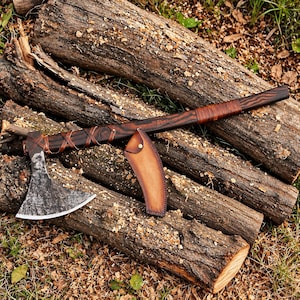 Può includere: Un'accetta con manico in legno scuro e testurizzato avvolto da un cordino in pelle marrone, dotata di una lama in metallo scuro usurato e un fodero in pelle color cuoio abbinato, fissato al manico con un bottone a pressione in ottone.