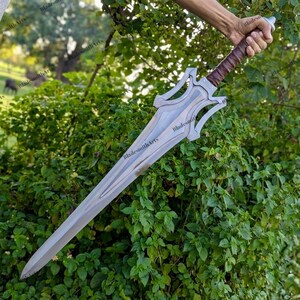 May include: A silver-colored metal sword featuring a double-edged blade with a central fuller, a complex open-work crossguard, and a brown leather-wrapped handle with a rounded pommel, held against a background of green leafy foliage.