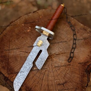May include: A short sword with a mottled silver-toned blade featuring a rectangular cutout and a gold-colored emblem. The handle is wrapped in brown leather with a silver-toned guard and a black metal chain attached to the pommel.