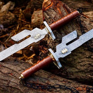May include: Two knives with textured silver-toned blades featuring a distinct cutout notch, a small gold-toned emblem, a silver-toned crossguard, and a brown leather-wrapped handle. A black metal chain is attached to the pommel of each knife.