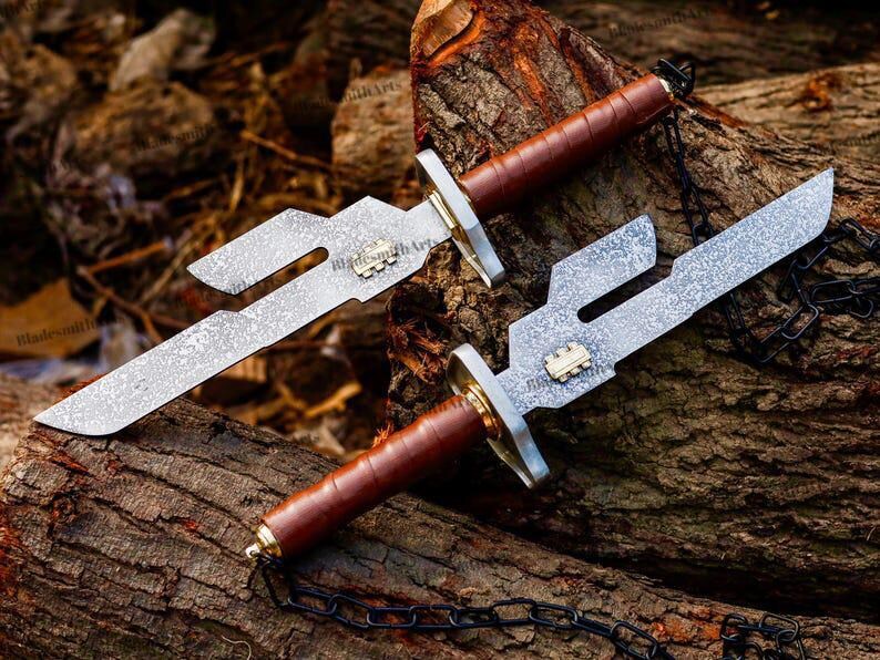 May include: Two knives with textured silver-toned blades featuring a distinct cutout notch, a small gold-toned emblem, a silver-toned crossguard, and a brown leather-wrapped handle. A black metal chain is attached to the pommel of each knife.