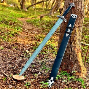 May include: A long, silver sword with a black handle and ornate silver accents, resting against a tree. The sword is paired with a black scabbard with silver detailing. The scene is set outdoors, with a forest backdrop.