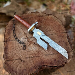 May include: A dagger with a textured silver-toned blade featuring a cutout section and a gold-colored inlay. The handle is wrapped in brown leather with a silver-toned guard and pommel. A black metal chain is attached to the pommel end.