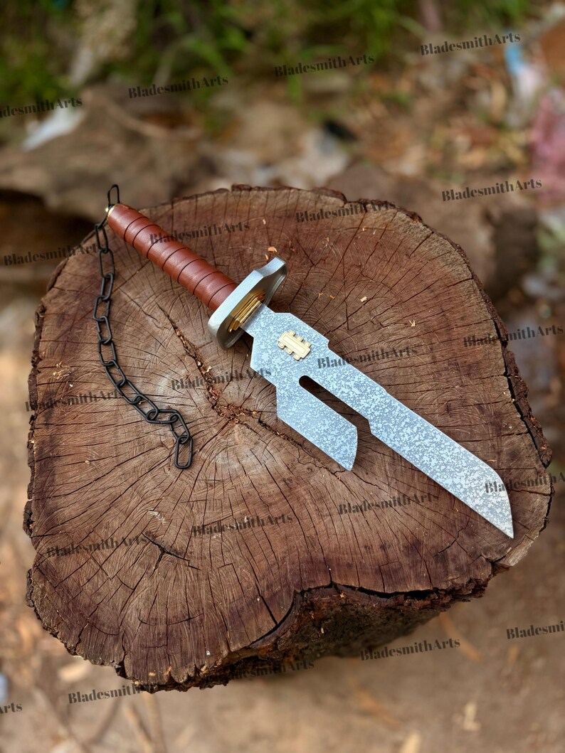 May include: A dagger with a textured silver-toned blade featuring a cutout section and a gold-colored inlay. The handle is wrapped in brown leather with a silver-toned guard and pommel. A black metal chain is attached to the pommel end.