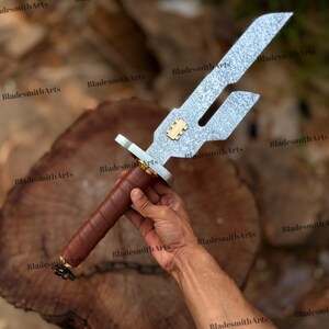 May include: A metal sword with a textured, silver-colored blade featuring a rectangular cutout and a gold-colored emblem. The handle is wrapped in brown leather, and a small black chain is attached to the pommel at the base of the grip.