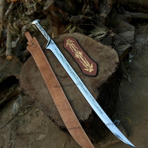 May include: A silver-colored metal sword with an etched blade and a patterned handle, accompanied by a long tan leather sheath and a separate dark brown leather piece featuring gold-colored decorative embroidery on its surface.