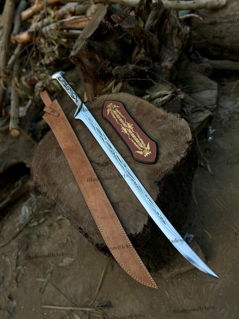 May include: A silver-colored metal sword with an etched blade and a patterned handle, accompanied by a long tan leather sheath and a separate dark brown leather piece featuring gold-colored decorative embroidery on its surface.