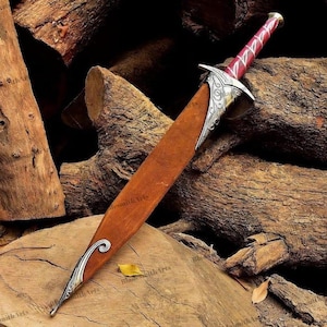 May include: A dagger with a brown leather sheath featuring silver-toned metal accents at the tip and hilt. The hilt has a deep red handle with silver-toned decorative engravings and a pommel. The metal components display intricate scrollwork patterns.