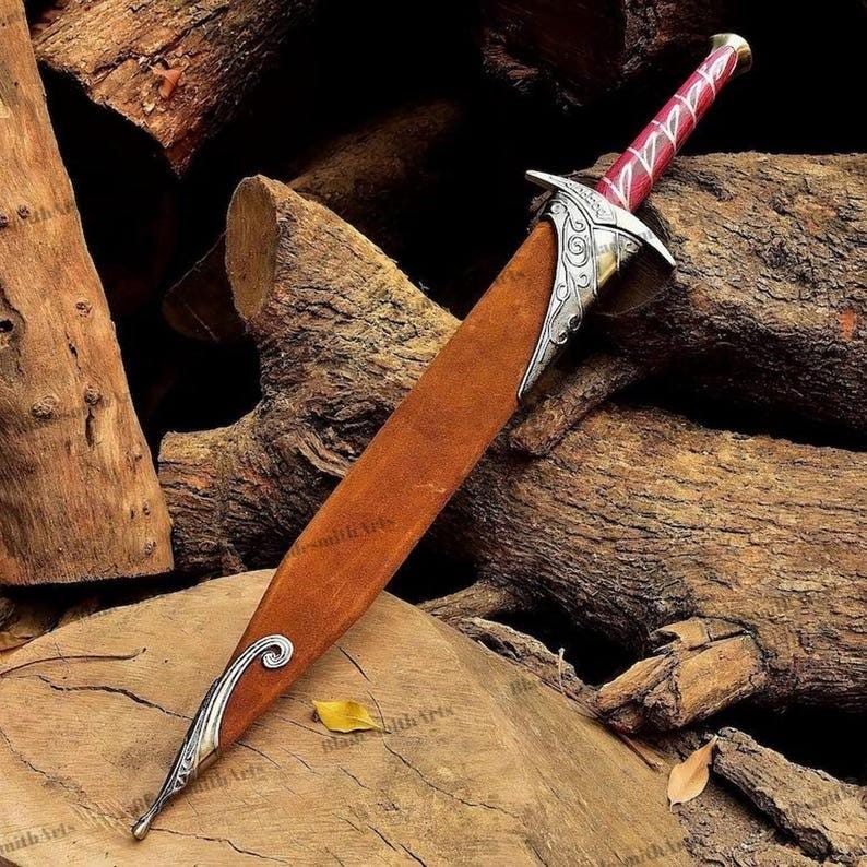 May include: A dagger with a brown leather sheath featuring silver-toned metal accents at the tip and hilt. The hilt has a deep red handle with silver-toned decorative engravings and a pommel. The metal components display intricate scrollwork patterns.