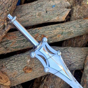 May include: A silver-colored metal dagger featuring a smooth cylindrical handle and a wide, pointed blade. The crossguard has a distinct geometric design with four rounded rivets at the corners, creating a symmetrical appearance with polished surfaces.