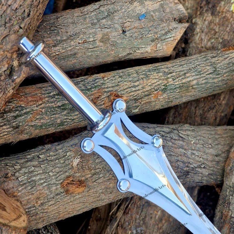 May include: A silver-colored metal dagger featuring a smooth cylindrical handle and a wide, pointed blade. The crossguard has a distinct geometric design with four rounded rivets at the corners, creating a symmetrical appearance with polished surfaces.
