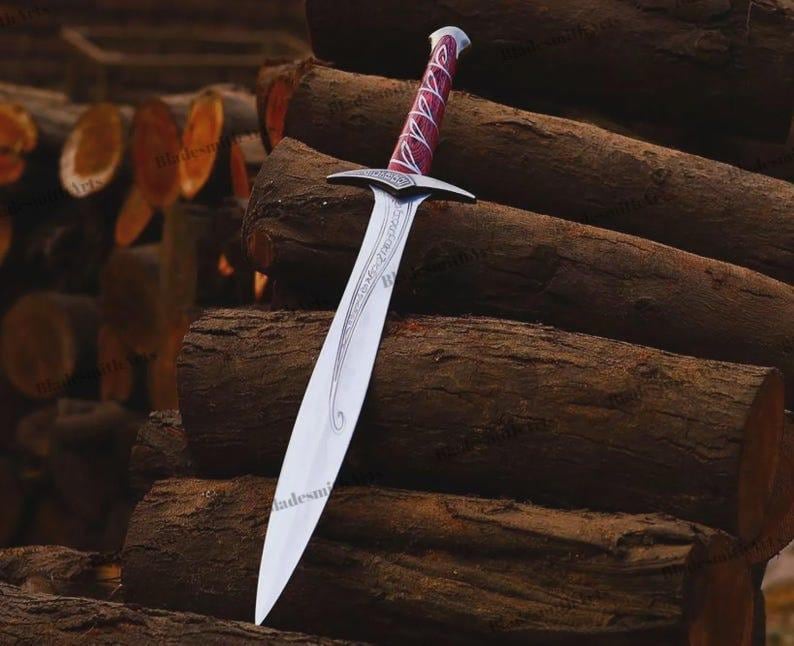 May include: A silver-toned metal sword featuring a pointed blade with intricate etched scrollwork patterns. The hilt has a metallic crossguard and a handle wrapped in a deep red material with a silver-colored pommel at the very end of the grip.