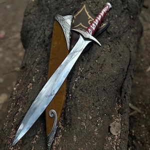 May include: A metal sword with a pointed blade featuring etched script, a silver crossguard, and a red handle with silver accents. The sword is accompanied by a brown scabbard with silver metal fittings at the tip and throat, displaying ornate scrollwork.