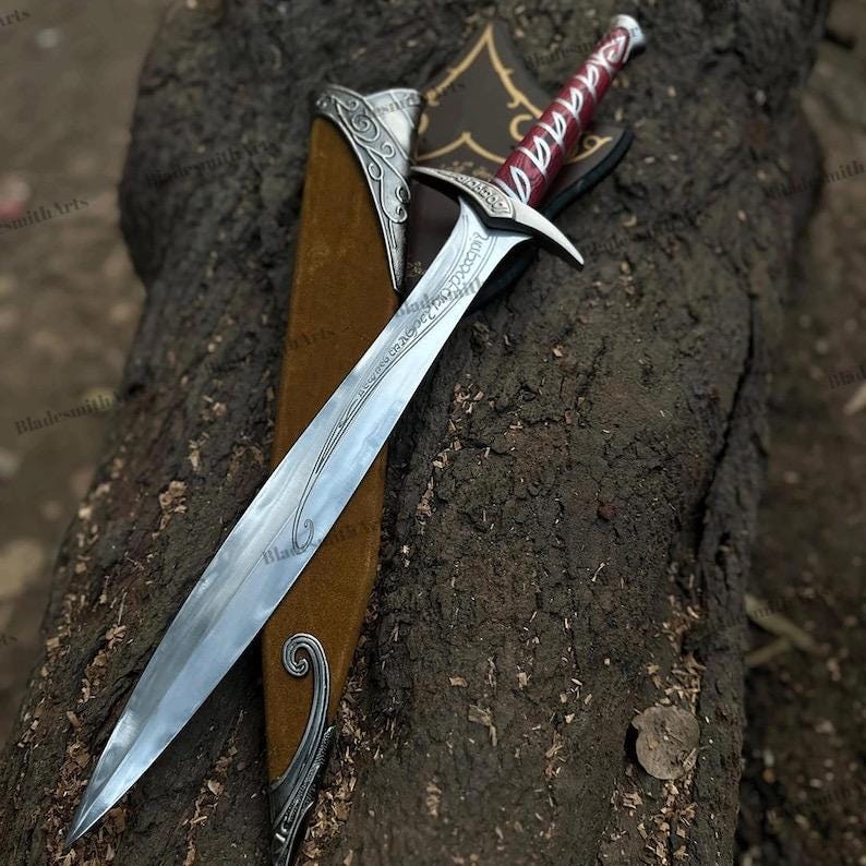 May include: A metal sword with a pointed blade featuring etched script, a silver crossguard, and a red handle with silver accents. The sword is accompanied by a brown scabbard with silver metal fittings at the tip and throat, displaying ornate scrollwork.