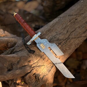 May include: A fixed-blade knife with a brown leather-wrapped handle, a gold-colored pommel and guard, and a patterned steel blade featuring a unique cutout design. A small gold-colored rectangular inlay is set into the blade near the guard.