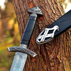 May include: A detailed image of a sword with a black handle and a silver-colored guard and pommel. The blade has a patterned design. The sword is next to a black sheath with a silver-colored decorative piece. The sword is leaning against a tree trunk.