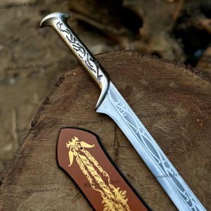 May include: A silver-toned metal sword with an intricate engraved handle and a blade featuring a flowing, vine-like pattern. Beside the blade lies a brown leather sheath decorated with a gold-colored, ornate floral design and the text BladesmithArts.