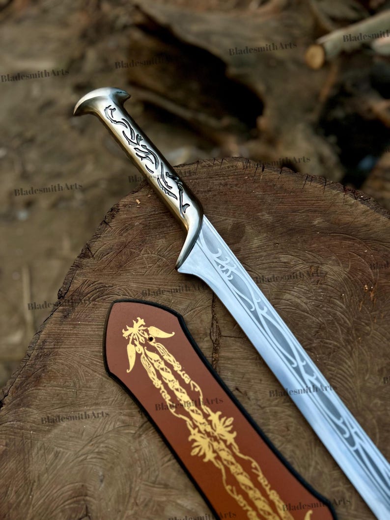 May include: A silver-toned metal sword with an intricate engraved handle and a blade featuring a flowing, vine-like pattern. Beside the blade lies a brown leather sheath decorated with a gold-colored, ornate floral design and the text BladesmithArts.