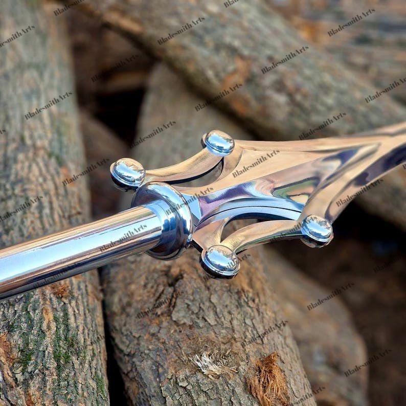 May include: A polished silver-toned metal spearhead featuring a central pointed blade flanked by two curved, wing-like protrusions. The design includes four small, rounded decorative rivets at the base of the blade and a cylindrical shaft.