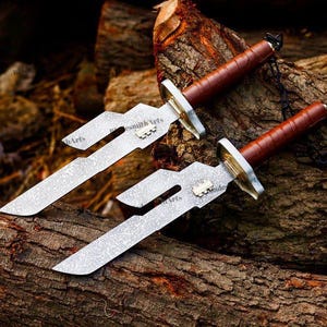 May include: Two knives with textured silver-toned blades featuring a distinct cutout design and a small metallic emblem. Each knife has a brown leather-wrapped handle, a metallic guard, and a black chain attached to the pommel end.