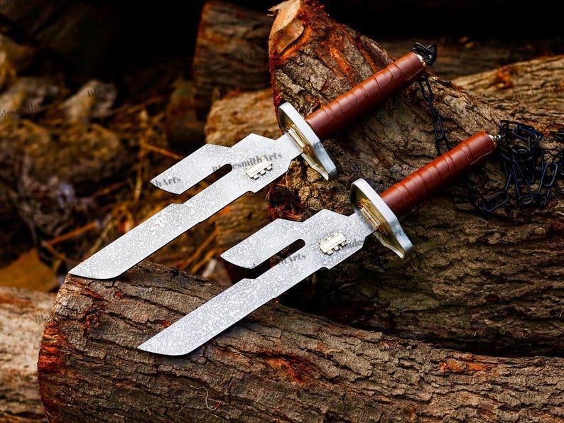 May include: Two knives with textured silver-toned blades featuring a distinct cutout design and a small metallic emblem. Each knife has a brown leather-wrapped handle, a metallic guard, and a black chain attached to the pommel end.