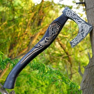 Pode incluir: Um machado preto com entalhes decorativos prateados. A cabeça e o cabo do machado apresentam desenhos intrincados. O machado é exibido ao ar livre, encostado a uma árvore, com folhagem verde ao fundo. O texto "Bladesmith Arts" é visível.