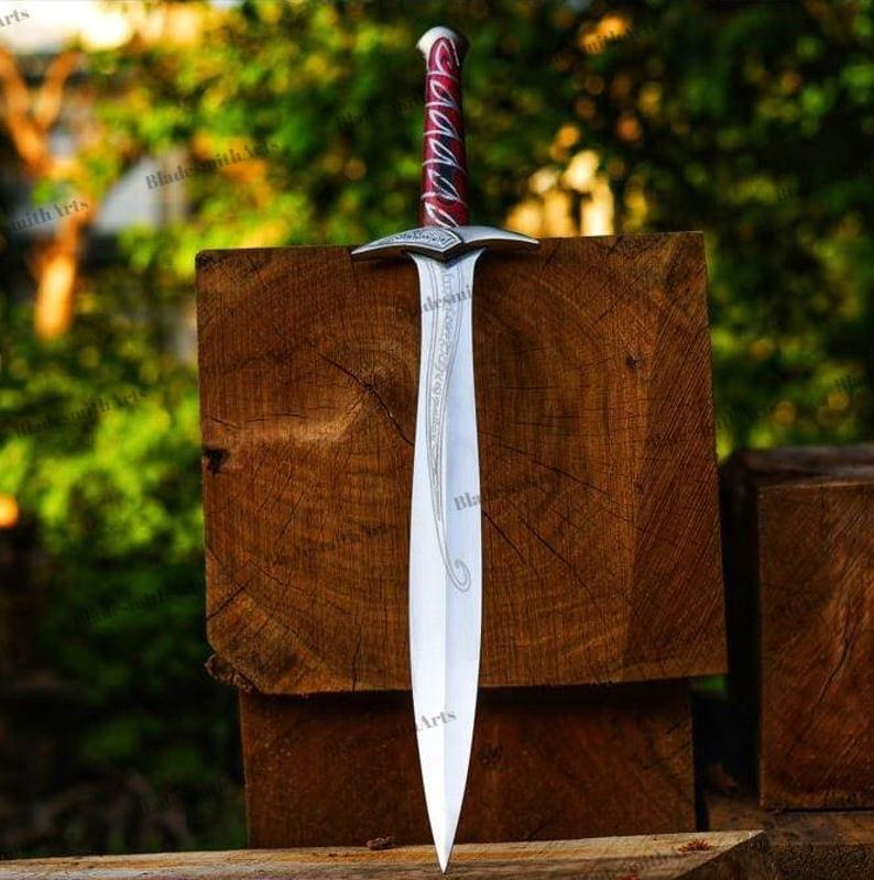 May include: A double-edged sword with a polished silver blade featuring intricate etched scrollwork. The hilt has a deep red grip wrapped in silver wire, a silver crossguard, and a rounded pommel. The blade tapers to a sharp point.