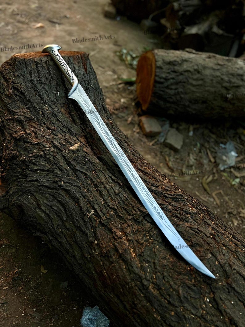 May include: A silver-toned metal sword featuring an elongated, curved blade with intricate, etched vine-like patterns running along the center and handle. The hilt has a rounded pommel and a pointed guard, with the text BladesmithArts visible.