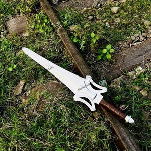 May include: A silver-colored metal sword with a pointed blade and a complex, cut-out crossguard design. The handle is wrapped in brown material, and the pommel is a rounded silver cap. The blade features the text Blade Arts printed on it.