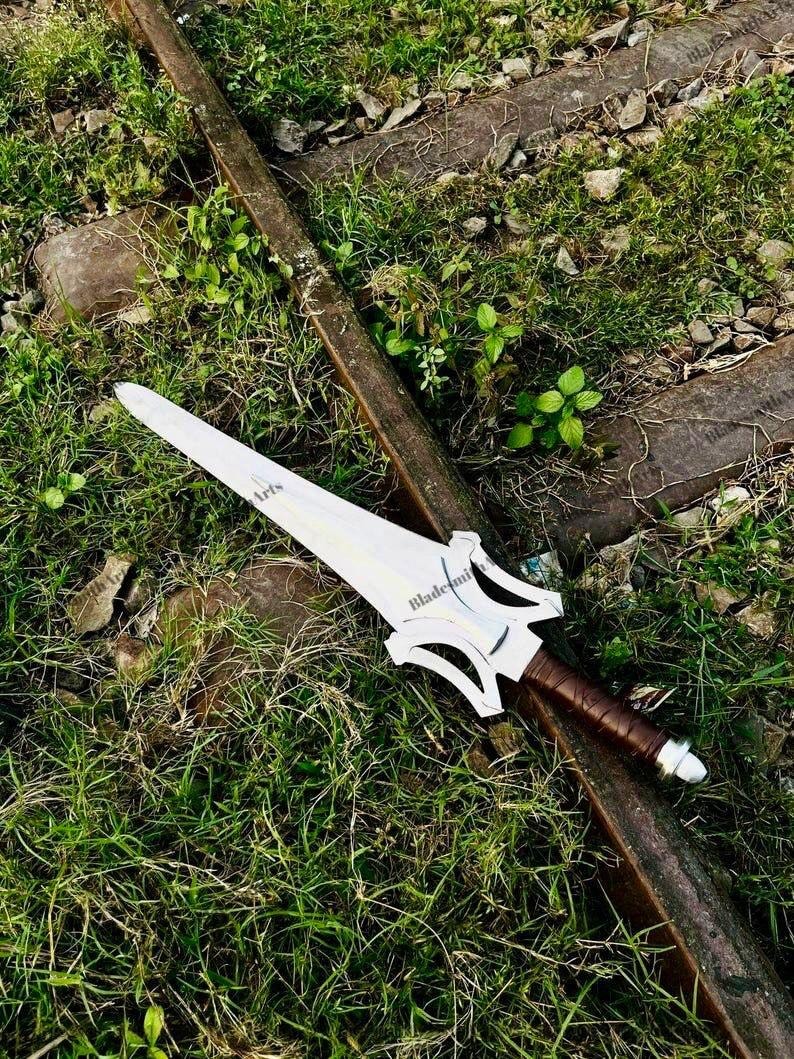 May include: A silver-colored metal sword with a pointed blade and a complex, cut-out crossguard design. The handle is wrapped in brown material, and the pommel is a rounded silver cap. The blade features the text Blade Arts printed on it.