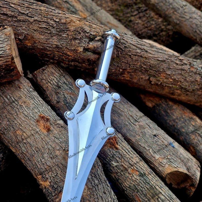 May include: A silver-colored metal dagger featuring a polished finish, a cylindrical handle, and a blade with a complex, multi-pointed guard design. The blade surface displays the repeating text BladesmithArts in a dark, subtle font.