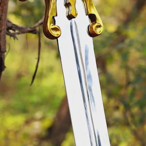 May include: A sword with a polished silver-colored metal blade featuring a central fuller groove. The crossguard is made of a shiny gold-colored metal with curved, scroll-like ends extending outward from the center of the hilt.