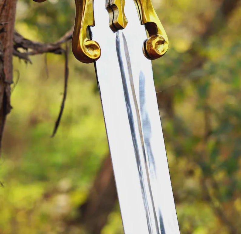 May include: A sword with a polished silver-colored metal blade featuring a central fuller groove. The crossguard is made of a shiny gold-colored metal with curved, scroll-like ends extending outward from the center of the hilt.