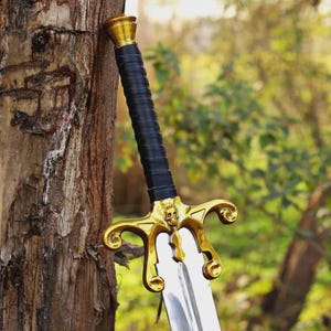 May include: A sword with a polished silver blade and a black leather-wrapped handle. The hilt features a gold-colored crossguard with ornate, curved ends and a central skull detail, topped with a matching gold-colored pommel cap.