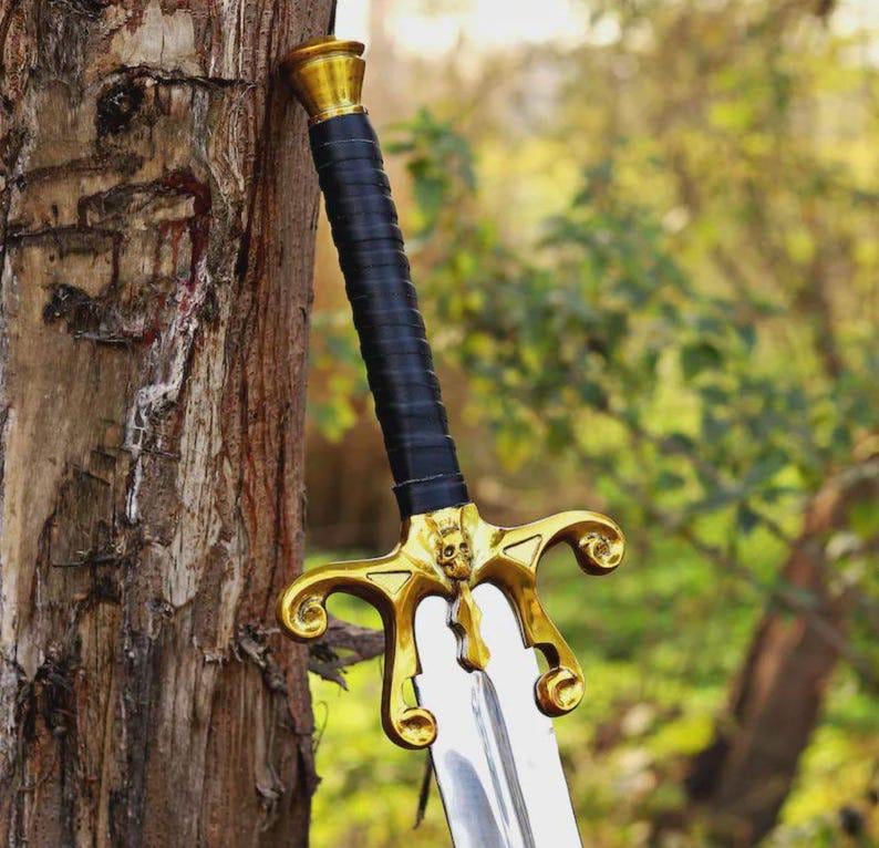 May include: A sword with a polished silver blade and a black leather-wrapped handle. The hilt features a gold-colored crossguard with ornate, curved ends and a central skull detail, topped with a matching gold-colored pommel cap.