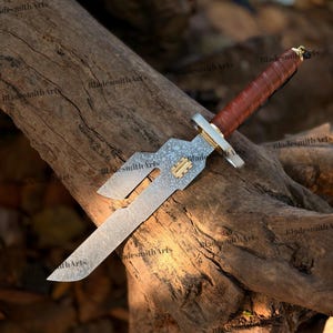 May include: A fixed-blade knife with a patterned steel blade featuring a unique cutout design and a small gold-colored rectangular inlay. The handle is wrapped in brown leather with a gold-colored guard and a small metal ring at the pommel.