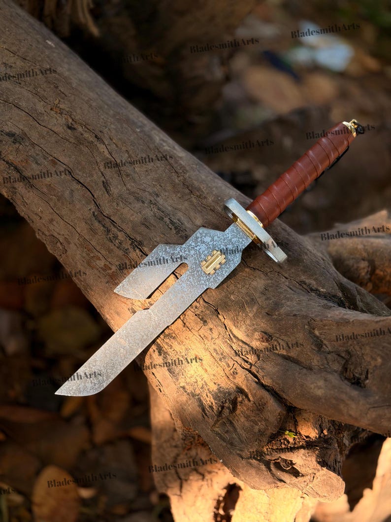 May include: A fixed-blade knife with a patterned steel blade featuring a unique cutout design and a small gold-colored rectangular inlay. The handle is wrapped in brown leather with a gold-colored guard and a small metal ring at the pommel.