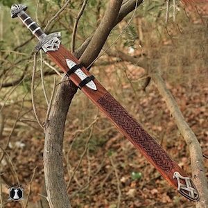 May include: A wooden sword with a brown sheath and intricate Celtic knotwork design. The sword features a silver-colored hilt and a decorative pommel. The sheath is secured with black straps, and the blade is not visible.