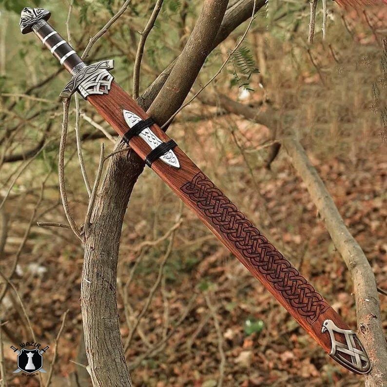 May include: A wooden sword with a brown sheath and intricate Celtic knotwork design. The sword features a silver-colored hilt and a decorative pommel. The sheath is secured with black straps, and the blade is not visible.