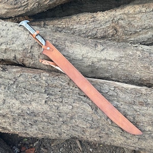 May include: A long, straight sword with a silver-toned metal hilt featuring engraved scrollwork details. The blade is encased in a tan leather scabbard that is secured with metal rivets and includes a thin leather strap attached to the side.