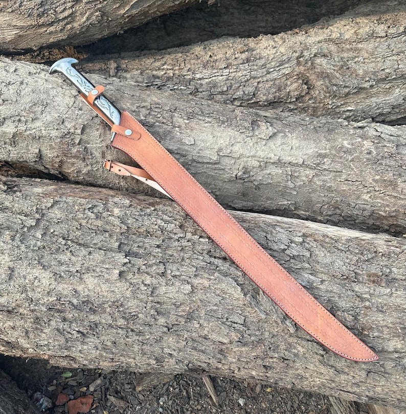 May include: A long, straight sword with a silver-toned metal hilt featuring engraved scrollwork details. The blade is encased in a tan leather scabbard that is secured with metal rivets and includes a thin leather strap attached to the side.