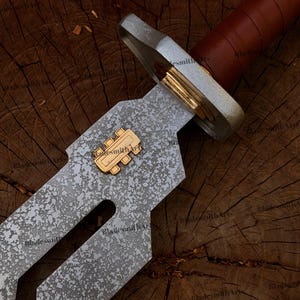 May include: A sword with a textured, mottled grey metal blade featuring a gold-colored rectangular emblem with three vertical bars. The hilt has a silver-toned crossguard, a gold-colored spacer, and a brown leather-wrapped handle.