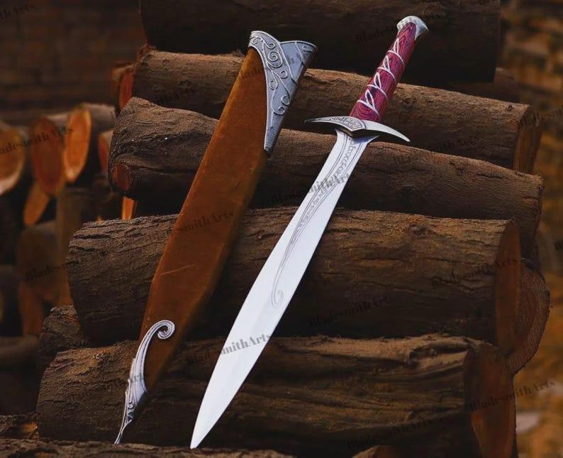May include: A silver-toned metal sword with a pointed blade and a purple-stained wooden handle featuring white decorative accents. The sword is accompanied by a brown sheath with silver-toned metal fittings at the top and bottom ends.