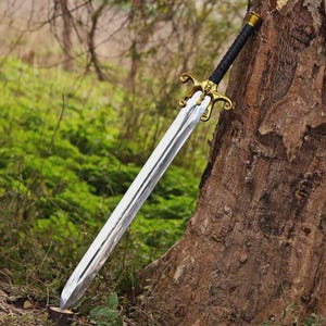 May include: A long sword with a polished silver-colored metal blade, a gold-toned metal crossguard featuring ornate curved details, and a black wrapped handle. The sword is positioned diagonally, resting against the textured bark of a tree trunk.