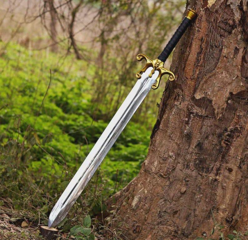 May include: A long sword with a polished silver-colored metal blade, a gold-toned metal crossguard featuring ornate curved details, and a black wrapped handle. The sword is positioned diagonally, resting against the textured bark of a tree trunk.