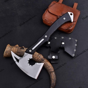 May include: A silver-colored metal axe head with a cutout gear-like center and a curved black handle. The axe is accompanied by a black leather sheath with silver rivets and a separate brown leather pouch, all arranged on a dark textured surface.