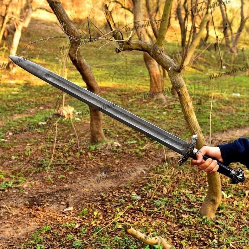 May include: A long, gray sword with a textured blade and ornate handle is held in a hand. The sword has a dark, metallic finish and a decorative crossguard. The handle is black with silver accents. The background features trees and foliage.