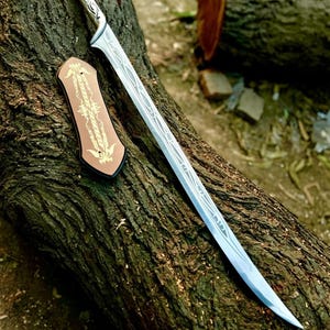 May include: A silver-toned metal sword with a curved blade featuring intricate etched line patterns and a matching handle. Beside the sword lies a small, rectangular, copper-colored plaque decorated with symmetrical gold-colored filigree designs.