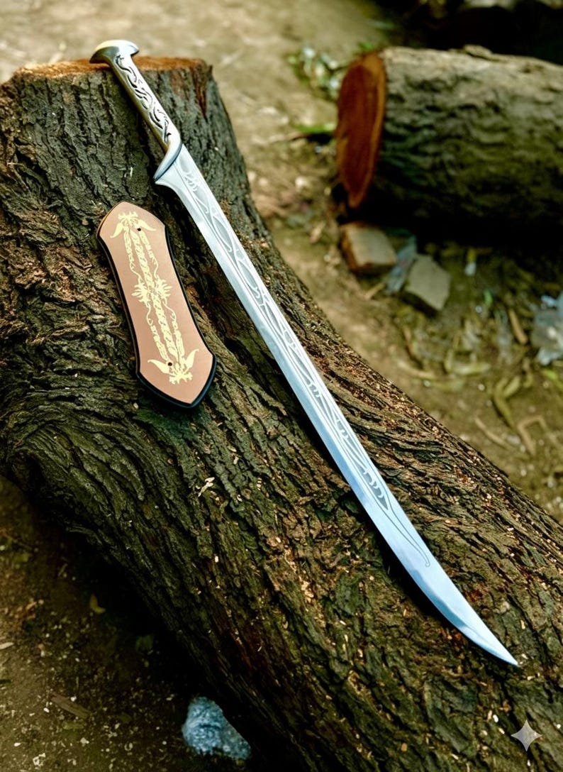 May include: A silver-toned metal sword with a curved blade featuring intricate etched line patterns and a matching handle. Beside the sword lies a small, rectangular, copper-colored plaque decorated with symmetrical gold-colored filigree designs.